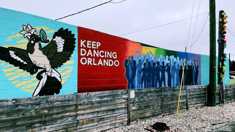 A mural on a brick wall with a lower wood wall in front of it. The mural shows a bird on the left with a flower in its beak. In the middle there is a rainbow gradient, the text “Keep Dancing Orlando”, and 49 human outlines to memorialize the 49 victims of the Pulse Club shooting