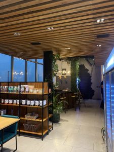 View larger photo: Modern café interior with wooden slat ceiling, large windows, and a shelving unit filled with various packaged items. The café features indoor plants and a counter area with high stools.