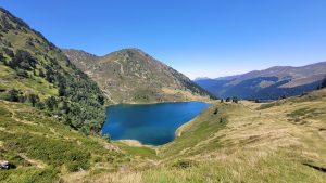 A picturesque mountain lake surrounded by lush green hills and forests under a clear blue sky. 