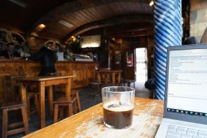 A close-up of a glass of dark coffee on a wooden table in a cozy café with rustic wooden furniture and a laptop open in the foreground displaying text on the screen.