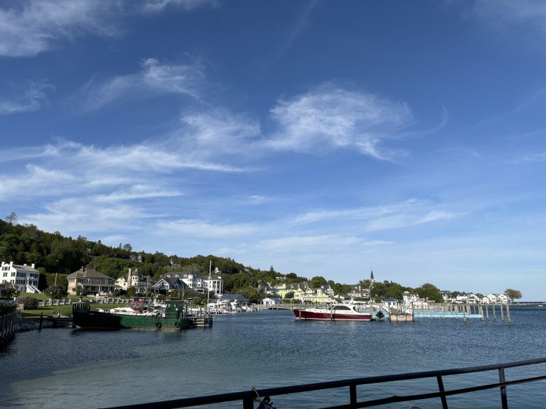 Mackinac Island Harbor, filled with boats, light clouds in a blue sky with the green island rising behind.