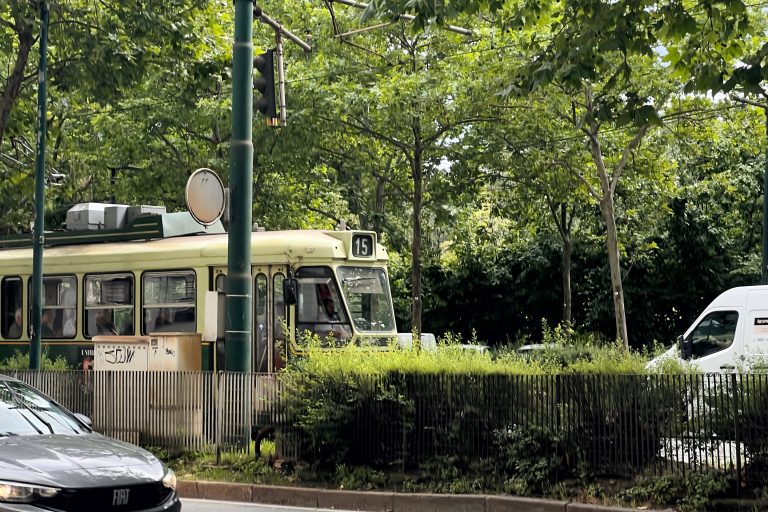 A vintage green tram labeled with the number 15 is stopped at a station surrounded by greenery, including trees and bushes. A black Fiat car and a white van are visible near the tram and there is a streetlight with a traffic light overhead.