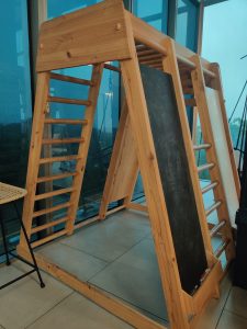 A wooden indoor play structure with climbing ladders, a blackboard with scribbles, and a shelf holding some chalk. The structure is placed on a tiled floor near large glass windows.