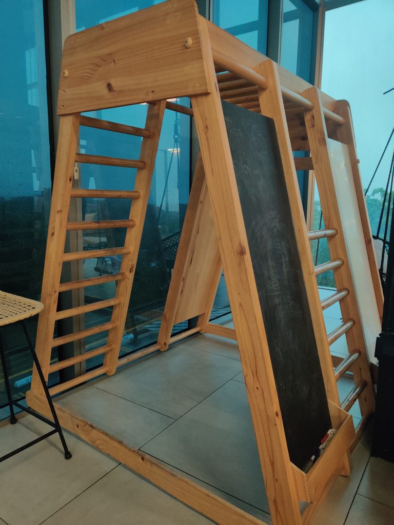 A wooden indoor play structure with climbing ladders, a blackboard with scribbles, and a shelf holding some chalk. The structure is placed on a tiled floor near large glass windows.