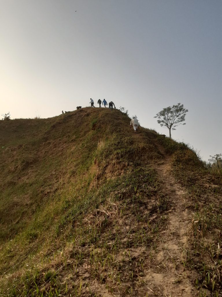 A group of people stand on top of a grassy hill while one person ascends the path toward them under a clear sky