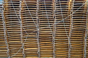 A large stack of wooden chairs piled on top of each other, secured with chains. The chairs are organized in rows and columns, creating a dense, grid-like appearance.