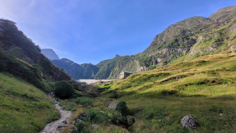 A scenic landscape of a mountain valley on a bright sunny day. The valley is lush with green grass, patches of bushes, and a narrow dirt path leading through it. A stone building with a slanted roof is situated on the right side of the valley, surrounded by towering, rugged mountains that are partially covered with green vegetation and rocky outcrops.