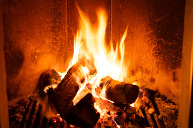 A close-up image of a lit fireplace. Burning logs are placed in the fire, and the flames are bright yellow and orange, creating a warm and cozy atmosphere.