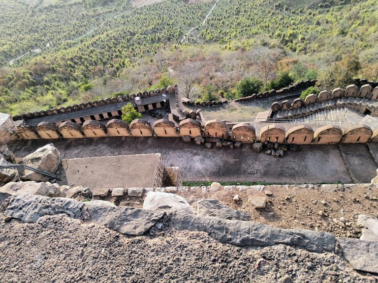 Aerial view looking down at the fortified walls of Kalinjar Fort, showcasing a series of arched structures lining the pathway. Beyond, dense green vegetation covers the surrounding hills.