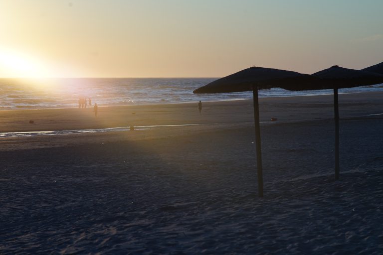 A serene beach at sunset, with the sun setting over the ocean, casting a warm glow on the water and sand. Few people can be seen in the distance, along with beach umbrellas in the foreground.