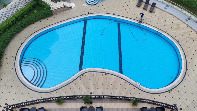 An aerial view of a large bean-shaped swimming pool with a person cleaning the deck on one side.