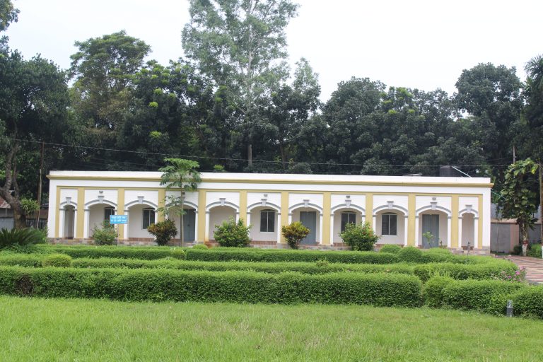 A single-story building with arched doorways and windows stands in front of a lush garden with trimmed hedges and green grass. The building is surrounded by tall, leafy trees.