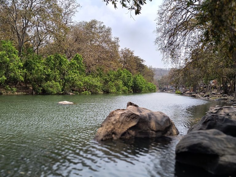 The calm waters of the Mandakini River reflect the vibrant green and autumnal foliage of the forest with few stones lining its banks under a clear blue sky.