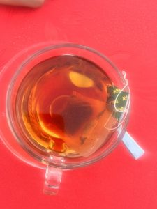 Looking down into a clear cup of black tea against a red tabletop. The teabag and its tag are to the right of the cup over the cup's handle.