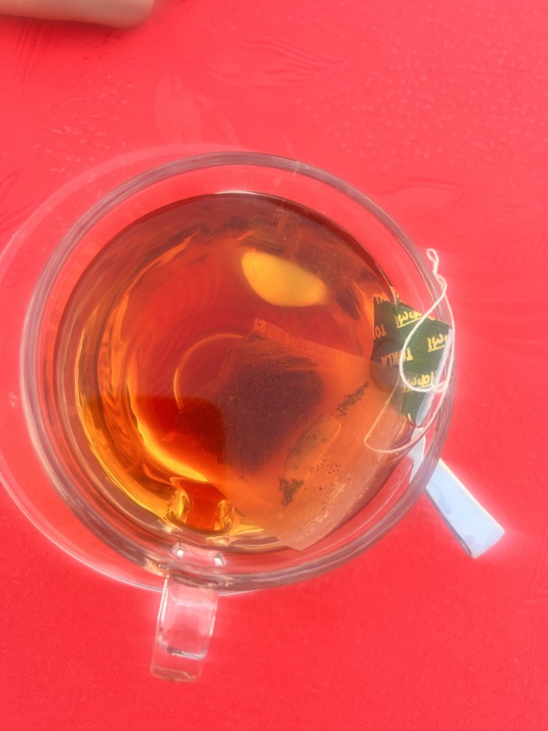 Looking down into a clear cup of black tea against a red tabletop. The teabag and its tag are to the right of the cup over the cup’s handle.