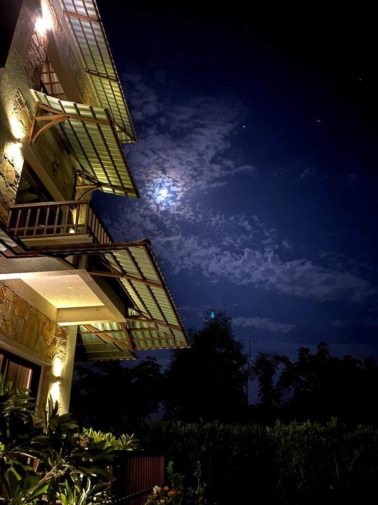 This image shows a nighttime view of a building with lighting. The sky is visible with scattered clouds partially covering the bright full moon.