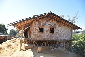 A traditional wooden stilt house with elevated bamboo walls and a thatched roof, located in a rural area with a dirt pathway and greenery in the background.