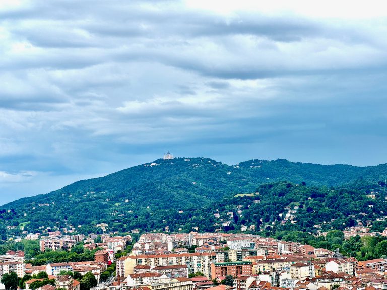 A view of a hillside in Torino, Italy, taken from the top of the Cinema Museum. The photo captures a densely forested hill with scattered buildings and the prominent Basilica of Superga (Basilica di Superga) at the top. The foreground features the cityscape with residential buildings and apartments. The sky is cloudy, adding a dramatic backdrop to