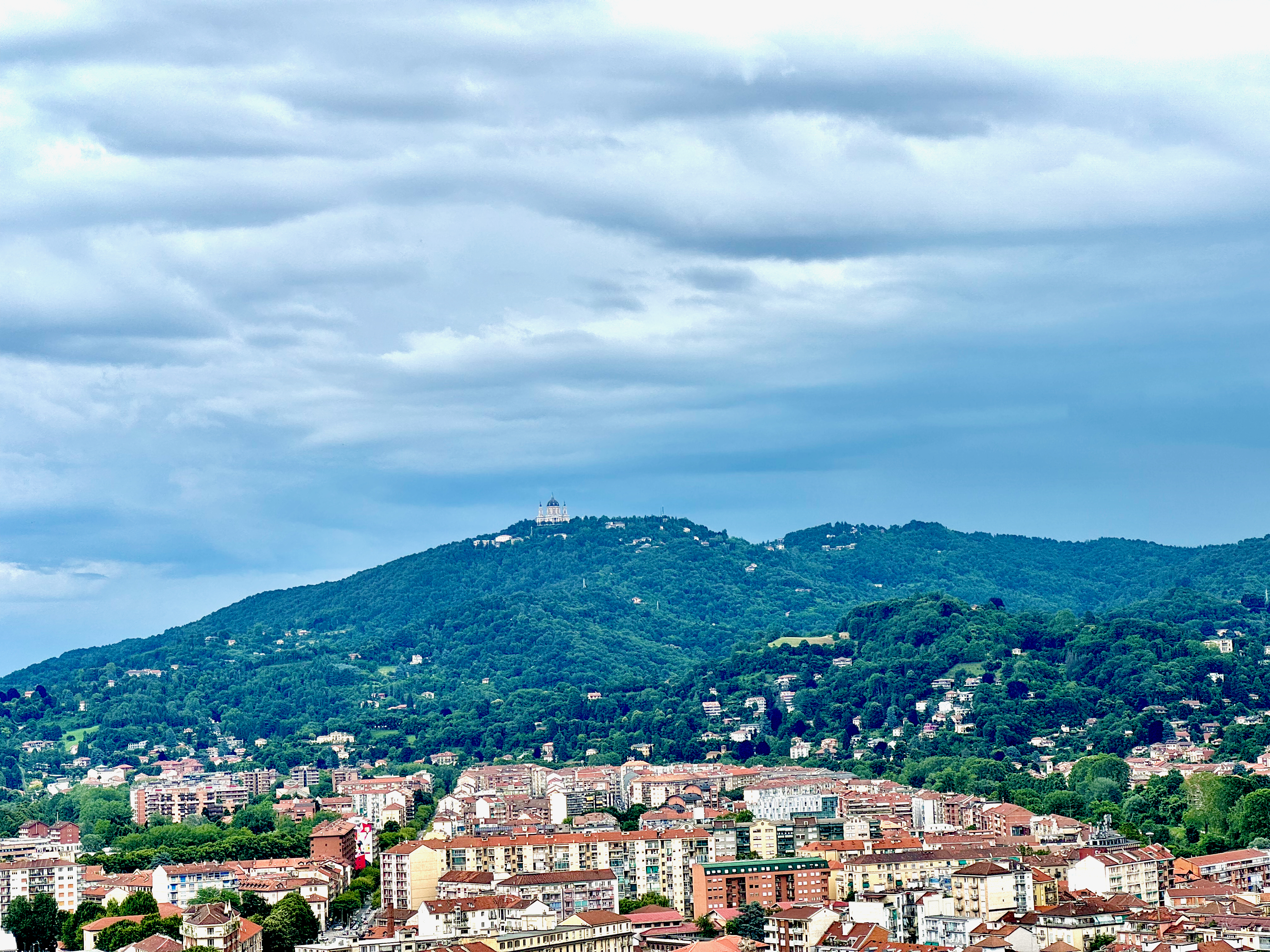 A view of a hillside in Torino, Italy, taken from the top of the Cinema Museum. The photo captures a densely forested hill with scattered buildings and the prominent Basilica of Superga (Basilica di Superga) at the top. The foreground features the cityscape with residential buildings and apartments. The sky is cloudy, adding a dramatic backdrop to 
