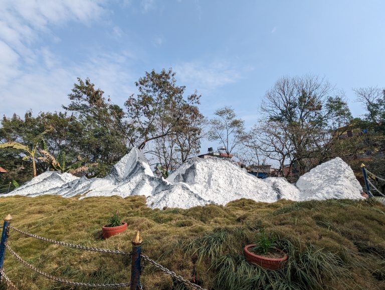 Artificial model mountain with a snow-capped design surrounded by grass and potted plants, situated in an outdoor area with trees and a clear sky in the background.