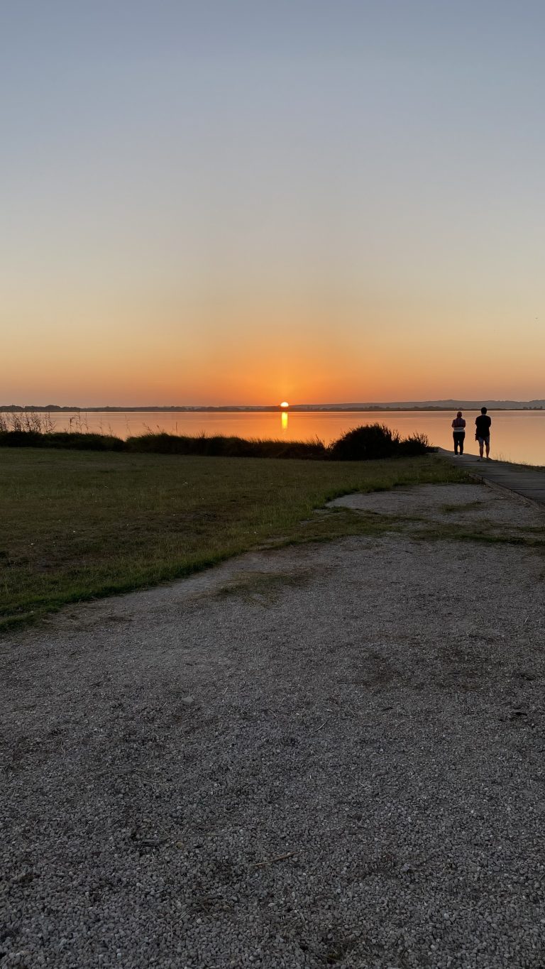 A serene scene of a sunset over a calm body of water. The sun is low on the horizon, casting a golden-orange glow across the sky and reflecting on the water’s surface. In the foreground, a gravel path leads to a grassy area with sparse vegetation. On the right side of the image, two people stand at the water’s edge, silhouetted against the sunset. The sky transitions from the warm hues near the horizon to a clear, pale blue higher up, indicating a clear and tranquil evening. The overall atmosphere is peaceful and contemplative.