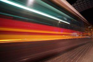 View larger photo: Long-exposure photo of a city street at night, capturing a vibrant, streaking blur of lights from a passing bus, predominantly featuring red, yellow, and teal colors.