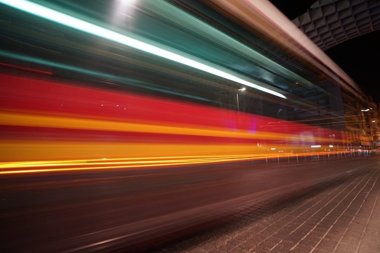 Long-exposure photo of a city street at night, capturing a vibrant, streaking blur of lights from a passing bus, predominantly featuring red, yellow, and teal colors.