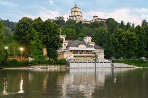 A view across the river Po in the city of Torino reveals a fairly large building used as a restaurant diagonally across the river, with the Chiesa di Santa Maria del Monte dei Cappuccini visible on top of a hill.