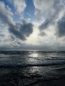 A serene seascape featuring the ocean with gentle waves reflecting the light of the sun partially obscured by dramatic, thick clouds in the sky.