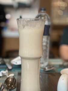 View larger photo: A tall glass filled with a creamy white beverage, standing on a table set with a spoon, a napkin, and a water bottle in the background.