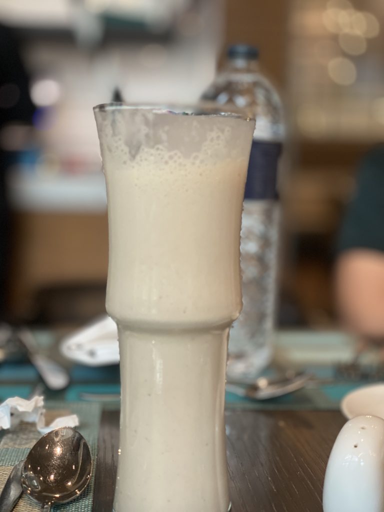 A tall glass filled with a creamy white beverage, standing on a table set with a spoon, a napkin, and a water bottle in the background.