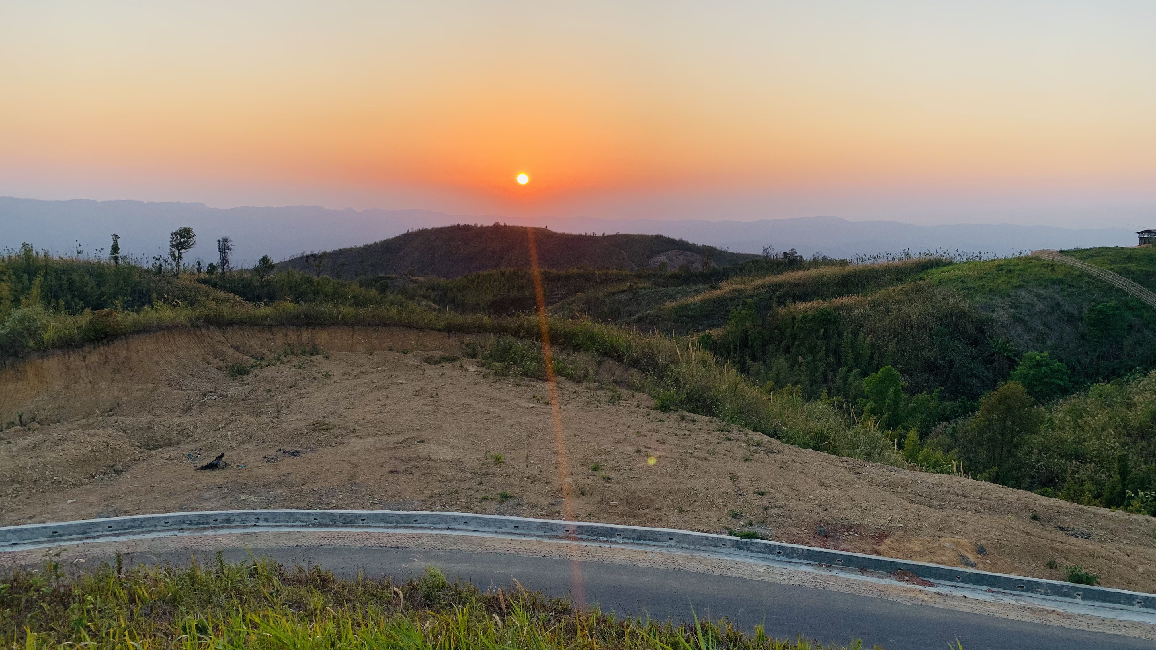 A scenic view of a sunset over rolling hills covered with vegetation. The sun is setting on the horizon, casting an orange and yellow glow across the sky. 