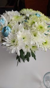 A bouquet of white and green fabric chrysanthemums, with a few blue flowers, arranged in a glass vase on a table.