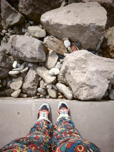 A person wearing brightly patterned pants and colorful shoes stands on a concrete surface near a collection of large and small rocks.