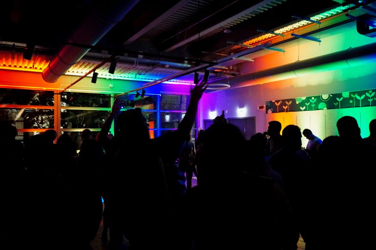 People dancing in a dark room illuminated by colorful lights in shades of red, green, and blue. The lights create a vibrant atmosphere, with the silhouettes of people clearly visible against the bright background.