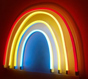 A brightly lit neon rainbow with red, orange, yellow, and blue colors arranged in an arching shape. The neon lights glow vividly against a dark background.
