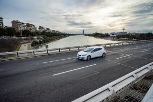 View larger photo: A white car is driving on an bridge across Guadalquivir river. Alongside the river runs a pedestrian path where two people are walking.