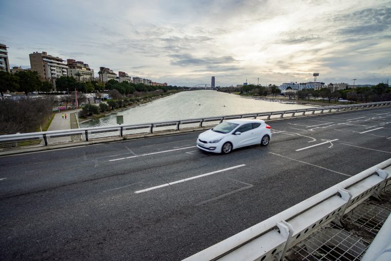 A white car is driving on an bridge across Guadalquivir river. Alongside the river runs a pedestrian path where two people are walking.