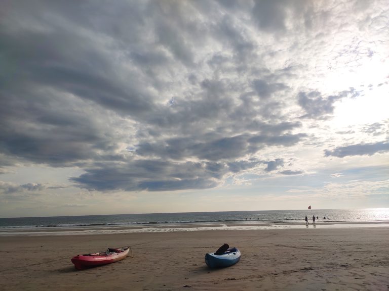 Tranquil evening at the beach, with kayaks resting on the shore and swimmers enjoying the calm sea under dramatic clouds.