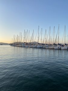 Sailboats lined up in a Sardinian marina with calm water in the foreground and a clear sunset or sunrise sky in the background.