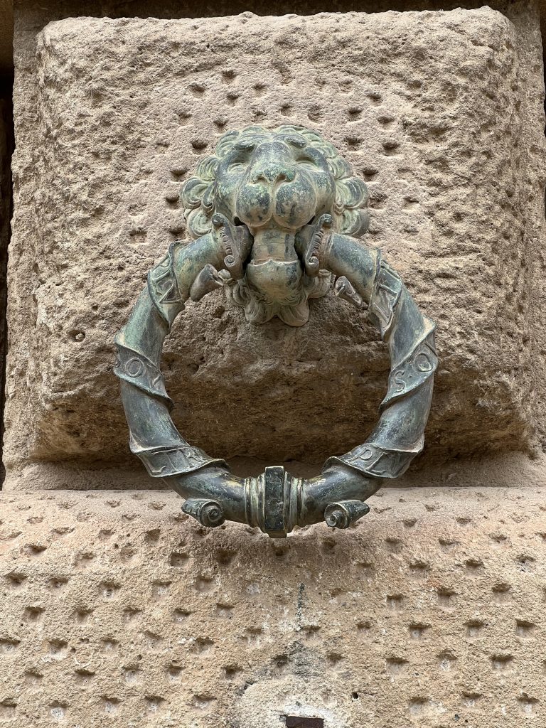 Metal ring attached to a wall with a lion’s head, also made of metal.