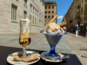  A photo of two desserts at the Gatsby Cafe in Torino, Italy. On the left is a tall glass of tiramisu espresso topped with whipped cream, served on a decorative plate with a spoon and napkin. On the right is a blue glass dish of hazelnut gelato garnished with two wafer rolls, also with a spoon and napkin. The background shows a sunny street.
