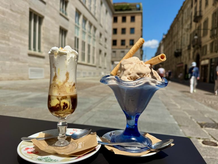 A photo of two desserts at the Gatsby Cafe in Torino, Italy. On the left is a tall glass of tiramisu espresso topped with whipped cream, served on a decorative plate with a spoon and napkin. On the right is a blue glass dish of hazelnut gelato garnished with two wafer rolls, also with a spoon and napkin. The background shows a sunny street.