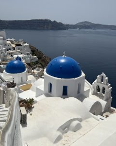 View larger photo: White buildings with iconic blue domes and a bell tower overlook a calm sea with distant cliffs and islands in the background, typical of Santorini, Greece.