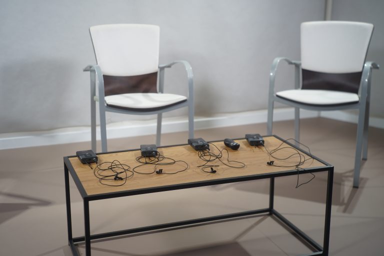 Two chairs with white and brown cushions positioned behind a wooden table, on which several lavalier microphones with cables are placed.