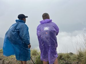 Two people standing outdoors, wearing blue and purple rain ponchos, looking at a foggy mountain landscape with grass and vegetation around them.