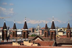 View larger photo: Multiple pointed brick towers topped with star ornaments, with a cityscape and snow-capped mountains in the background under a clear blue sky.