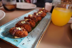 A plate of breaded croquettes topped with a wasabi and garnished with slices of fresh tuna and sesame seeds, served on a long blue dish. There is a glass of orange juice with ice beside the dish. 