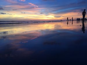 View larger photo: Sunset at Cox's Bazar, with bright yellow and orange colors reflecting on the beach's wet sand, and silhouettes of several people in the distance.