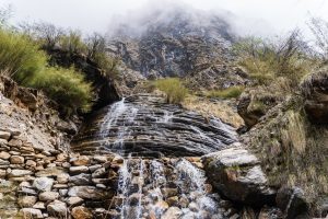 A small waterfall cascades over layered, rocky terrain, surrounded by sparse vegetation and shrubs. In the background, a steep, rugged mountain partially obscured by mist rises, adding a dramatic and mystical atmosphere to the scene. The foreground features a rocky stream bed with variously sized stones.